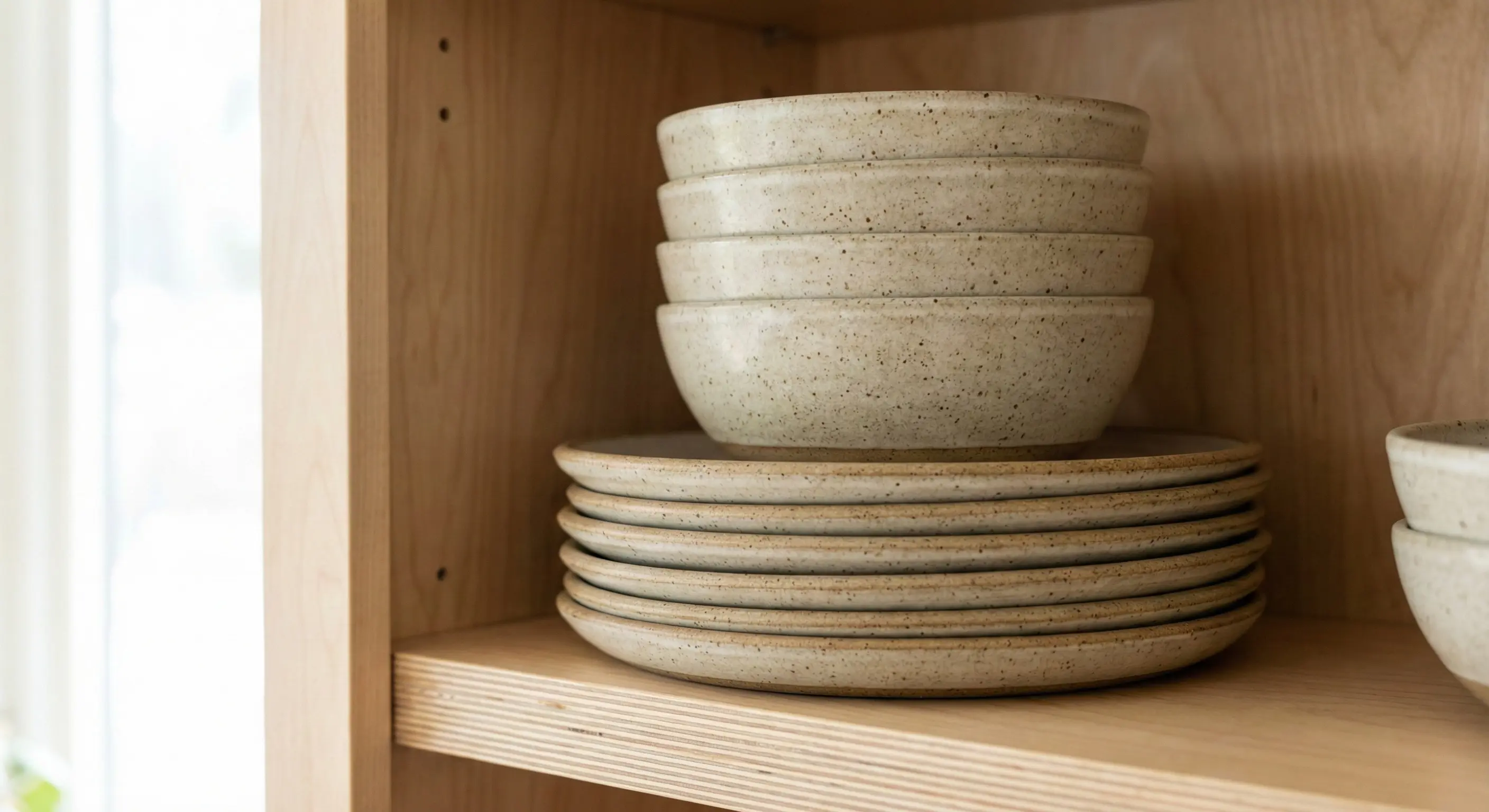 A neat stack of speckled ceramic stoneware plates and bowls on a wooden kitchen shelf, showing their thickness and matte glaze