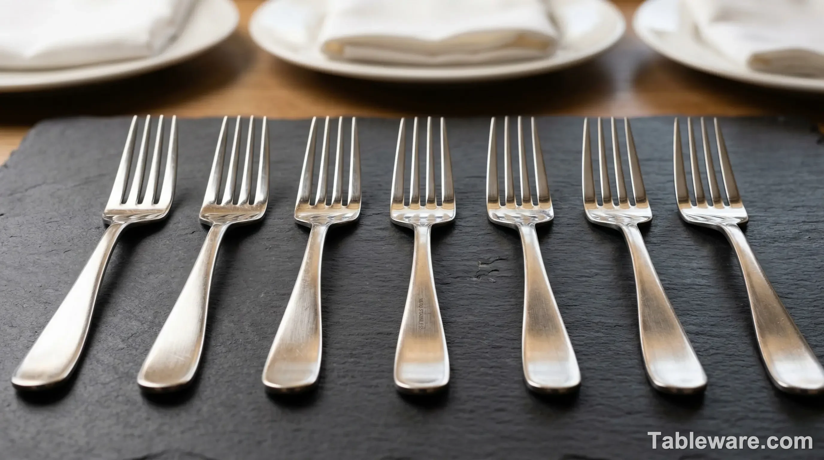 A minimalist close-up shot of high-quality 18/10 stainless steel forks resting on a smooth dark slate surface.