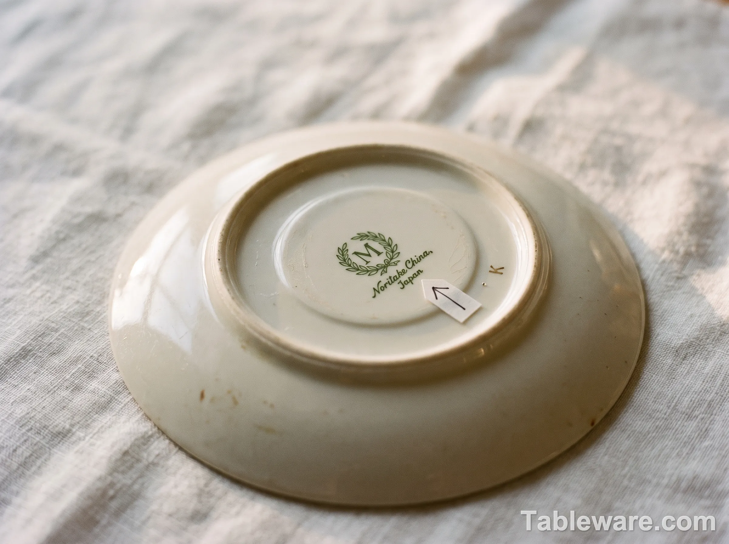Editorial photograph of a Noritake M-in-wreath back stamp on the bottom of a 1950s china saucer, with an arrow pointing to the dating letter code