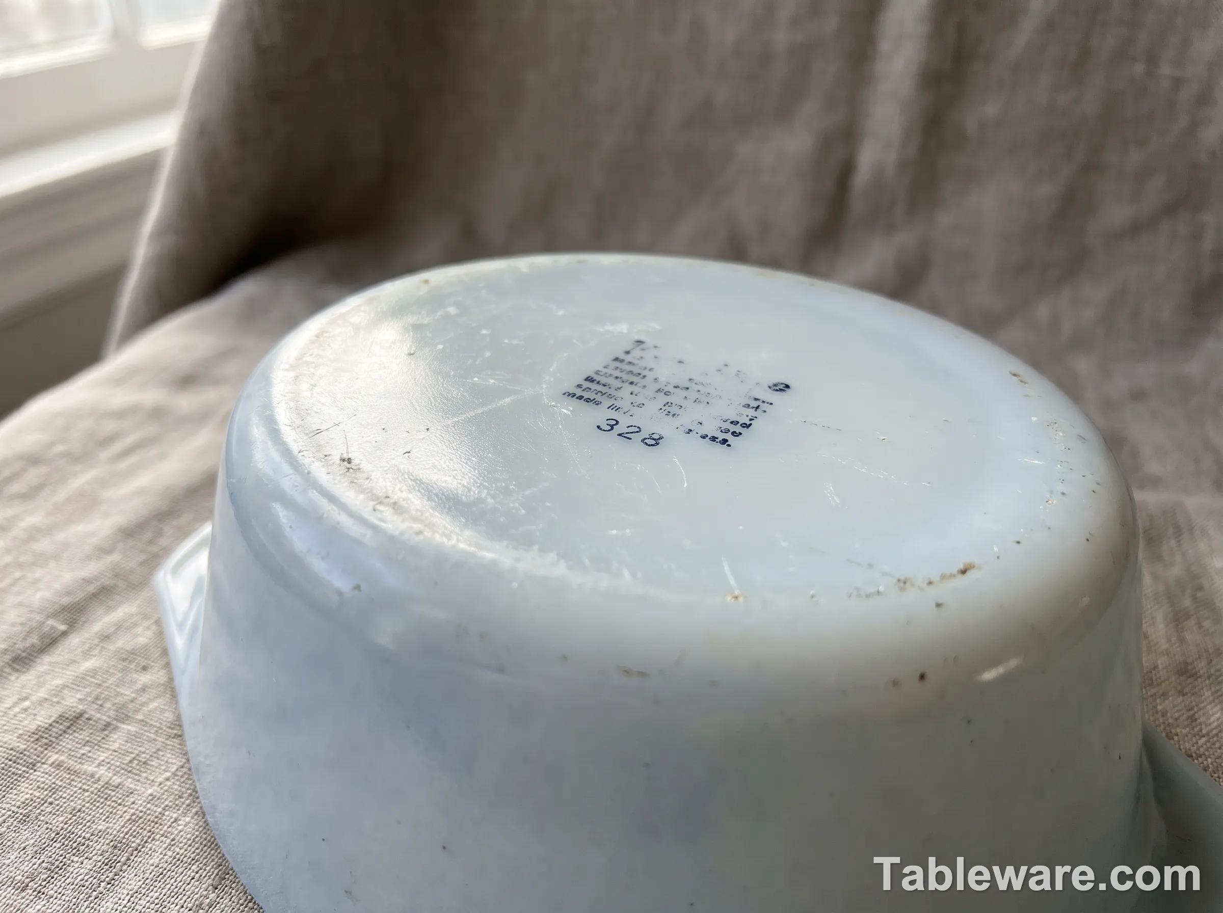 Macro view of the underside of a vintage milky white opal glass casserole dish resting on neutral linen, showing a faded stamped backstamp with printed lettering and a three digit model number, soft raking window light, aged glass patina